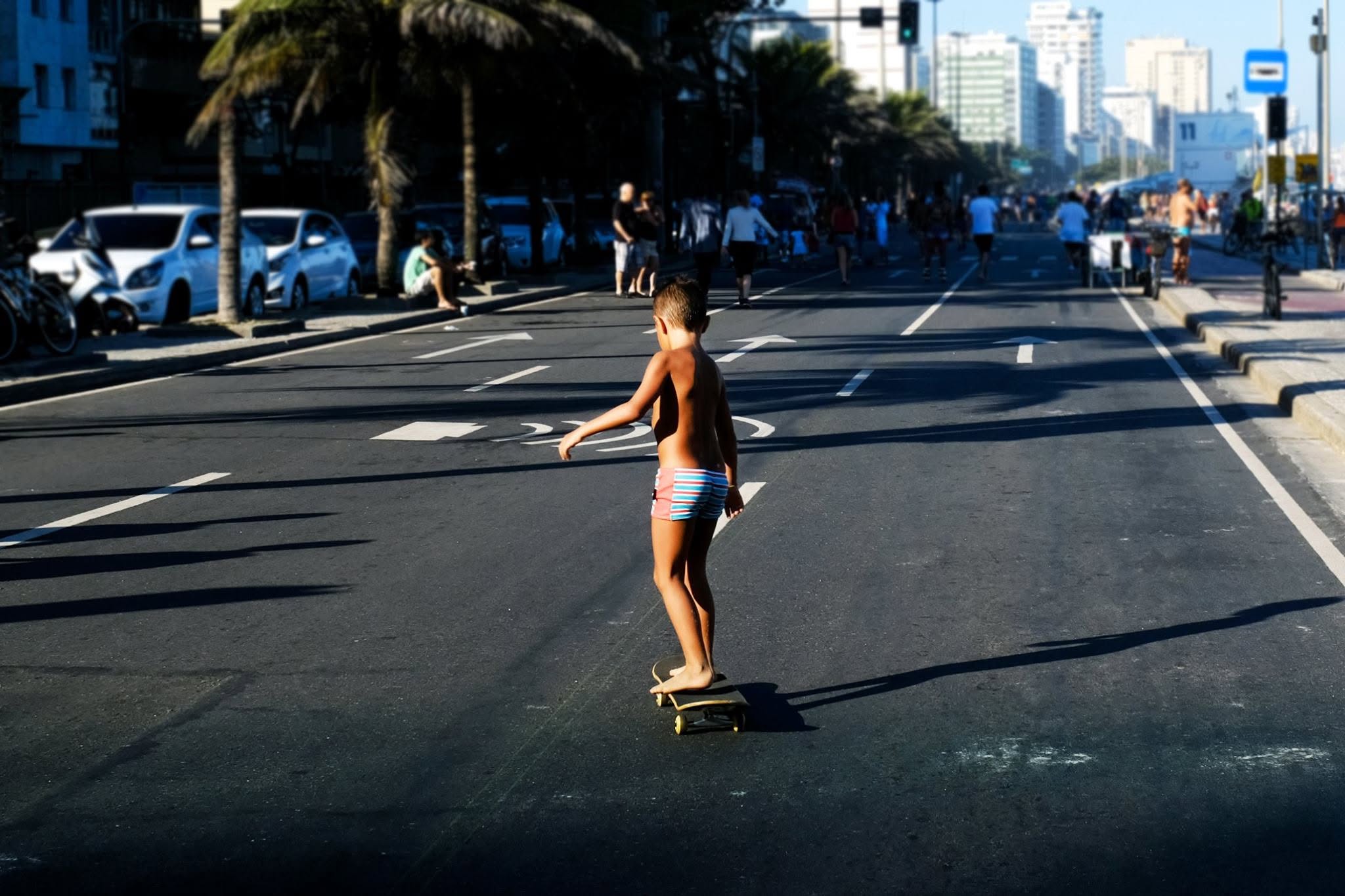 A young skateboarder rolls along a sunlit street in Rio de Janeiro, casting a long shadow.