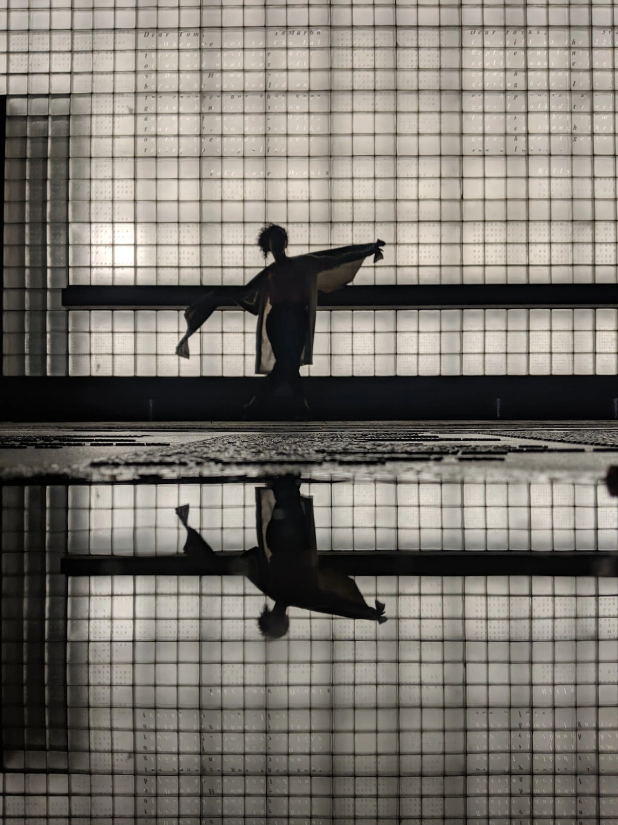 A vertical photograph of a silhouetted figure holding fabric and its reflection beneath the luminous, lettered memorial wall in Lower Manhattan.