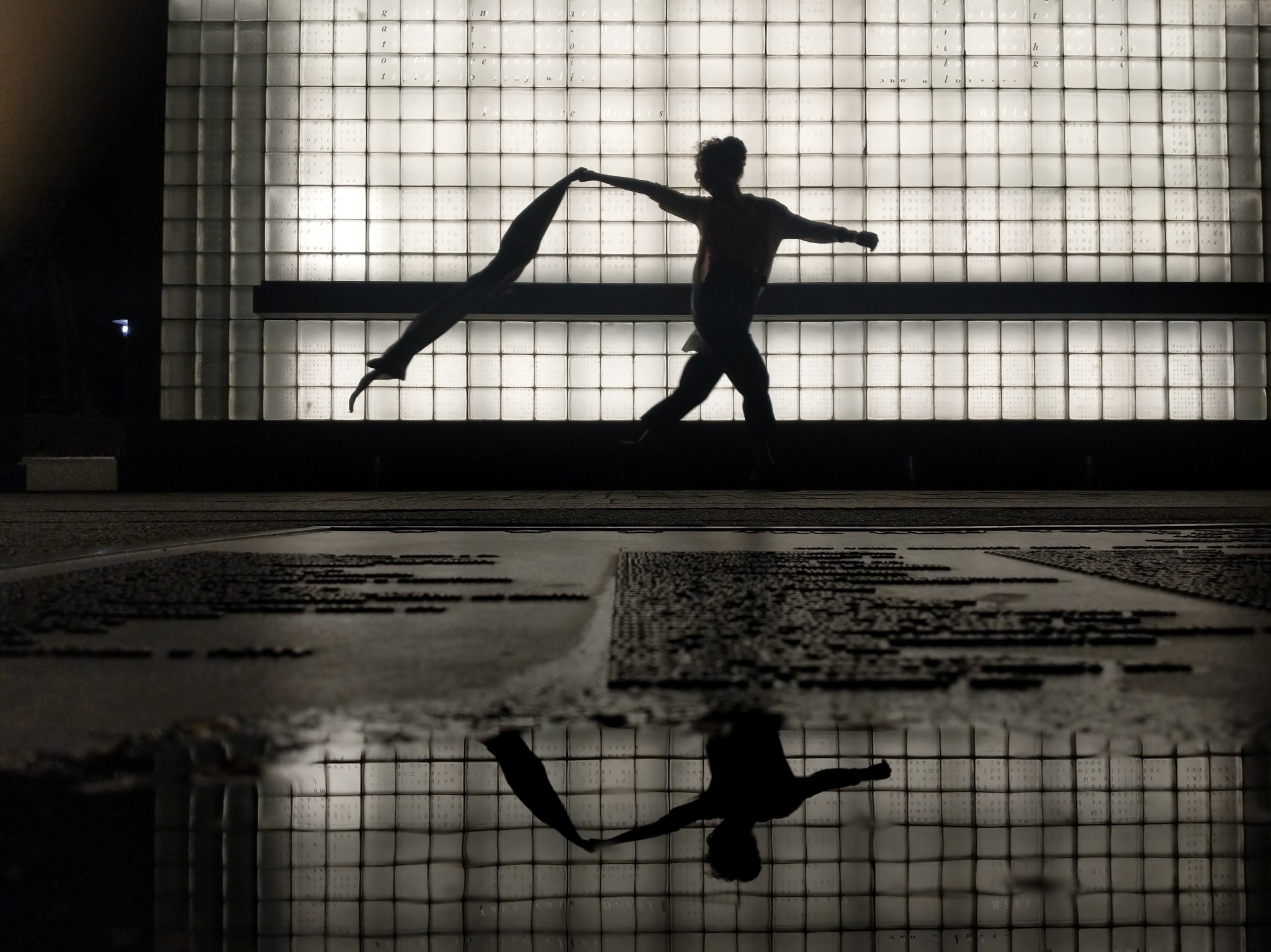 A silhouetted figure holding fabric is reflected below a luminous glass-block memorial wall in Lower Manhattan.